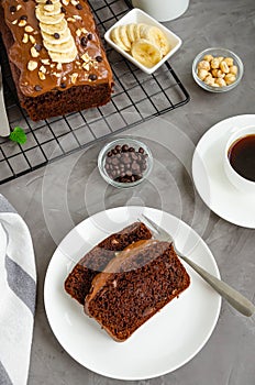 Homemade chocolate banana bread with chocolate cream, banana slices and nuts on top on a dark concrete background.