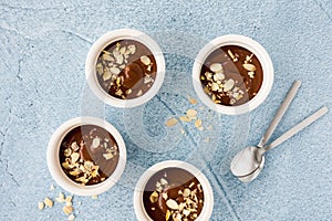 Homemade chocolate pudding in three white ceramic ramekins with roasted almond slivers and teaspoons on light blue concrete
