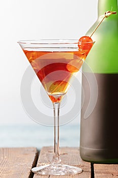 A homemade cocktail with cherries in a martini glass with a wine bottle behind on a wooden table