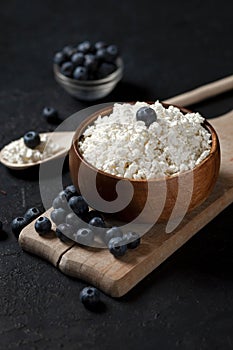 Homemade cottage cheese with blueberries in a wooden plate against a dark background, healthy milk breakfast with berries on a