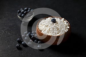 Homemade cottage cheese with blueberries in a wooden plate against a dark background, healthy milk breakfast with berries on a