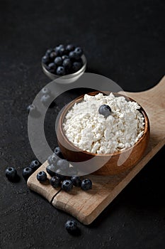 Homemade cottage cheese with blueberries in a wooden plate against a dark background, healthy milk breakfast with berries on a