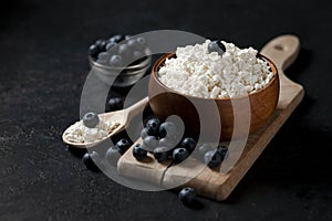 Homemade cottage cheese with blueberries in a wooden plate against a dark background, healthy milk breakfast with berries on a