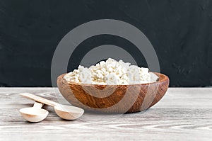 Homemade cottage cheese, curd in a bowl with two crossed spoons on a white rustic wooden table with black background.