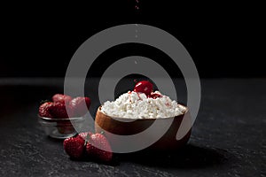 Homemade cottage cheese with strawberries and jam in a wooden plate against a dark background, adding red cream to a milk