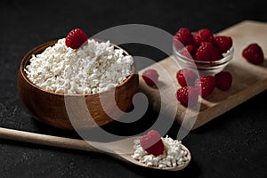 Homemade cottage cheese in a wooden plate with raspberries in a rustic bowl against a dark background, healthy food on a