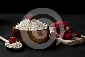 Homemade cottage cheese in a wooden plate with raspberries in a rustic bowl against a dark background, healthy food on a black