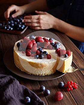 Homemade creamy mascarpone New York Cheesecake with berries on dark wooden table. Hands in the frame.