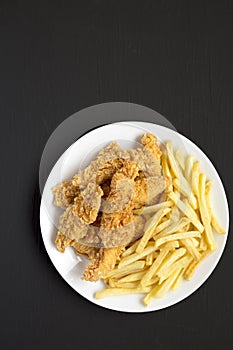 Homemade Crispy Chicken Tenders and French Fries on a white plate on a black surface, top view. Space for text