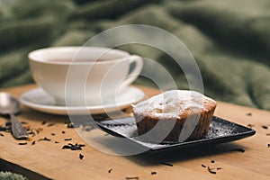 Homemade cupcake with powdered sugar on a black plate and a white cup of tea with natural additives on a wooden tray