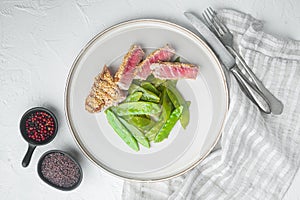 Homemade Cut Grilled Sesame Tuna steak with spring onions and sugar snap peas, on plate, on white stone  background, top view flat