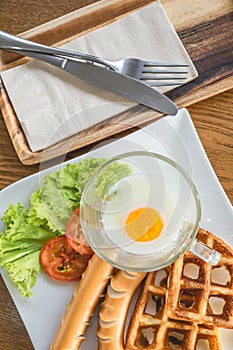 Homemade delicious american breakfast with soft-boiled egg, waffles, sausage, tomato, lettuce on white plate on wood table, top