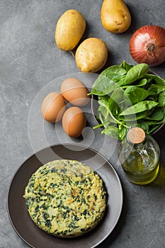 Homemade delicious frittata with spinach on plate. Recipe ingredients potatoes eggs olive oil in bottle on dark stone