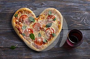 Homemade delicious margarita pizza with tomatoes and basil in the shape of a heart with a glass of red wine on a wooden table for