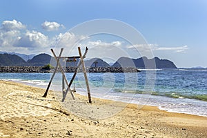 Homemade empty rope swing on a deserted beach against the background of the sea and blue sky with clouds.