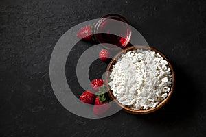 Homemade fresh cottage cheese without additives in a wooden plate with strawberries and red jam on a black texture table