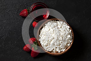 Homemade fresh cottage cheese without additives in a wooden plate with strawberries and red jam on a black texture table, top view