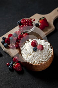 Homemade fresh cottage cheese with strawberries, raspberries, blueberries, currants in a wooden plate against a dark background,