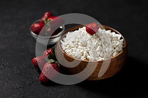 Homemade fresh cottage cheese in a wooden plate without additives lies with strawberries on a black texture table