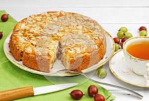 Homemade gooseberry pie garnished with almond petals and cup of tea on white wooden table.