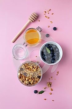 Homemade granola with nuts and seeds in jar, honey and blueberries on pink background. Granola for healthy breakfast.