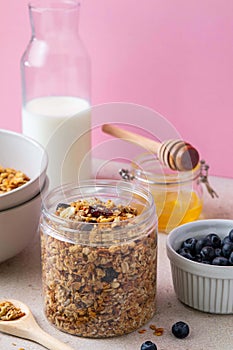 Homemade granola with raisin, seeds, peanut and hazelnut in glass jar, milk bottle, honey and blueberries on pink background.