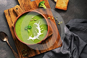 Homemade green cream soup of broccoli with sour cream and pumpkin seeds in a clay bowl on a dark background. Overhead view, flat