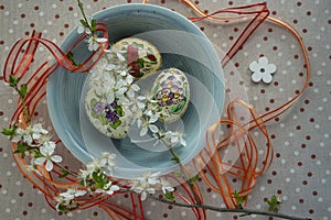 Homemade handmade painted Easter eggs in blue bowl on spotted tablecloth decorated with blackthorn sloe branch and red ribbon