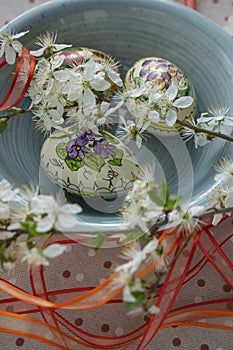 Homemade handmade painted Easter eggs in blue bowl on spotted tablecloth decorated with blackthorn sloe branch and red ribbon