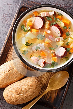 Homemade hearty pea soup with vegetables, bacon and sausages close-up in a bowl. Vertical top view