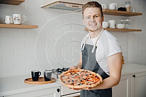 Homemade Italian pizza, handsome young male chef in dark apron holds baking sheet in bright kitchen. Cooking video blog