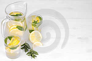 Homemade lemonade with lemon and mint in a glass jug and a glass next to lemon on a white wooden background