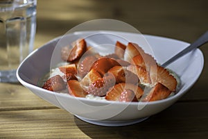 Homemade oatmeal porridge bowl with sliced strawberries and a glass of water on table in morning sunlight