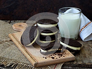 Homemade Oreo chocolate cookies with white marshmallow cream and glass of milk on dark background.