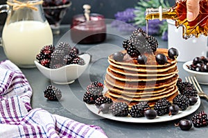 Homemade pancakes with blackberries and currants are stacked on a plate and watered with maple syrup, in the background berries