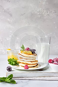 Homemade pancakes with jam, frozen raspberries, blackberries and mint and glass with milk on light grey background.