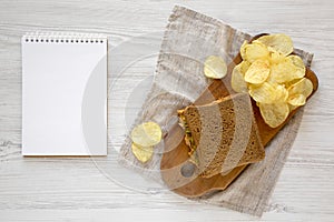 Homemade Peanut Butter Pickle Sandwich with Potato Chips on a rustic wooden board, blank notepad on a white wooden background, top
