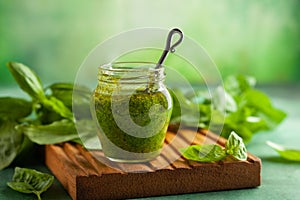 Homemade pesto sauce of fresh basil, parmesan and pine nut in glass jar with spoon on wooden board. Front view, copy space