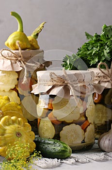 Homemade pickled patissons, cucumbers with carrots and garlic in glass jars and fresh ingredients on a light gray background,