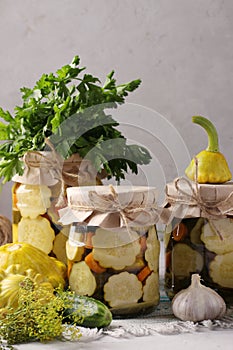 Homemade pickled patissons, cucumbers with carrots and garlic in glass jars and fresh ingredients on a light gray