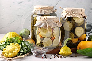 Homemade pickled patissons, cucumbers and zucchini in glass jars and fresh ingredients on a light gray background in