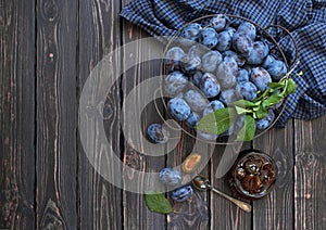 Homemade plum jam in a glass jar and fresh blue plums in a bowl on a dark rustic wooden background with copy space top view.