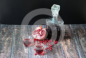 Homemade pomegranate liqueur in a bottle and two glasses and a broken fruit with a scattering of seeds on a wooden table