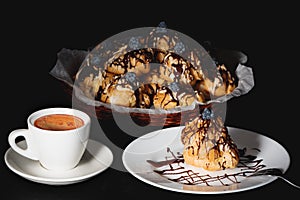 Homemade profiteroles with custard cream topped with dark and white melted chocolate, cup of coffee on black background