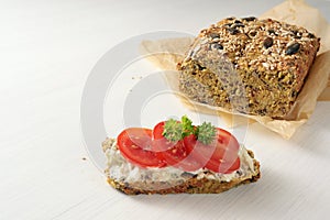 Homemade protein bread with seeds on baking paper and sandwich with curd, tomatoes and parsley garnish on a white table, healthy