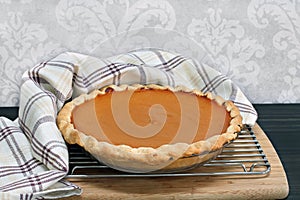 Homemade pumpkin pie on a cooling rack with towel wrap.