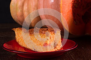 Homemade pumpkin pie for Thanksgiving on a red saucer, and a large fresh sliced orange pumpkin, wooden background, close