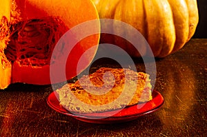 Homemade pumpkin pie for Thanksgiving on a red saucer, and a large fresh sliced orange pumpkin, wooden background, close