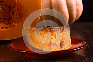 Homemade pumpkin pie for Thanksgiving on a red saucer, and a large fresh sliced orange pumpkin, wooden background, close