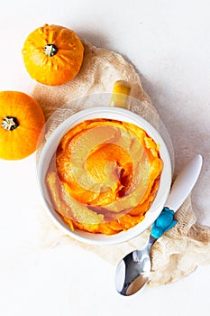 Homemade pumpkin puree in bowl with baby spoon and fresh pumpkins on light concrete background.